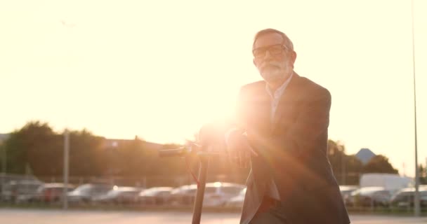 Portrait prise de vue du vieil homme caucasien en lunettes appuyé sur scooter électrique et regardant la caméra. Grand-père sénior aux cheveux gris dans un véhicule à l'extérieur. Homme pensionné en ville au coucher du soleil. - Séquence, vidéo