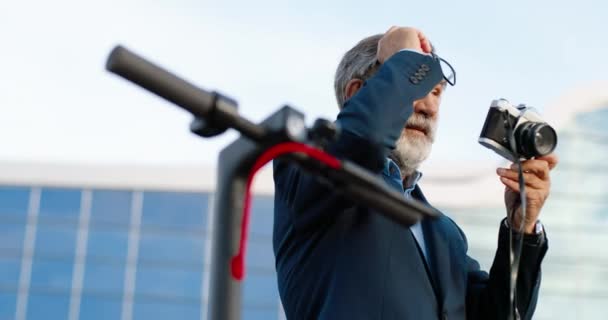 Кавказький старий сивоволосий чоловік у окулярах стоїть надворі і фотографує його фотокамерою. Велосипед або електричний скутер. Старший чоловік - фотограф, який працює на вулиці в міському середовищі.. - Кадри, відео