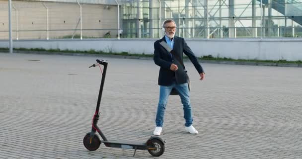 Hombre mayor en gafas y auriculares con bolsa de pie en scooter eléctrico en la calle urbana, escuchando música y bailando alegre y divertido. Viejo macho en airpods danza y moviéndose divertido fuera. - Metraje, vídeo