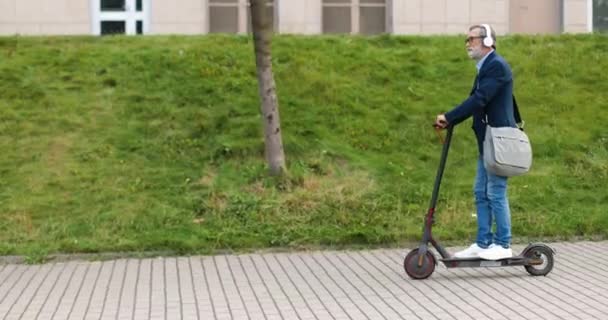 Beyaz saçlı, gözlüklü, kulaklıklı, elektrikli scooterın üzerinde duran ve bir yerlere giden yaşlı bir adam. Yaşlı erkek emekli ya da iş adamı şehir aracını kullanıyor ve açık havada müzik dinliyor.. - Video, Çekim