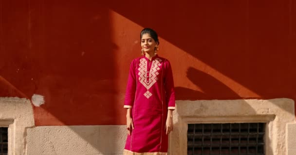 Portret hinduskiej młodej pięknej kobiety z kropką na czole i w czerwonym tradycyjnym stroju uśmiechniętym do aparatu szczęśliwie. Na zewnątrz. Dość uroczy uśmiechnął radosną szczęśliwą dziewczynę z Indii na czerwonej ścianie na ulicy - Materiał filmowy, wideo