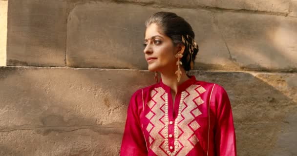 Retrato de la joven hindú hermosa mujer con punto en la frente y en traje tradicional rojo girando la cara y sonriendo a la cámara. Al aire libre. Bastante encantadora chica feliz de la India en la pared antigua en la calle - Metraje, vídeo