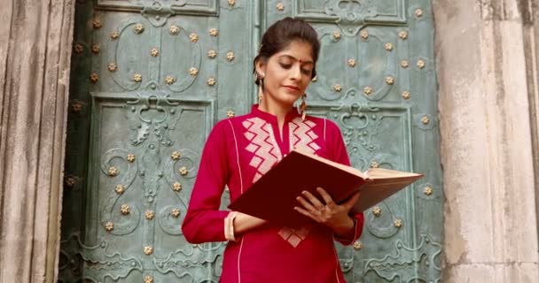 Portret van een prachtige jonge Hindoe vrouw met stip op het voorhoofd die buiten voor de deur staat en glimlacht naar de camera. Leesboek. Mooie stijlvolle charmante vrouw in rode traditionele outfit met leerboek. - Video
