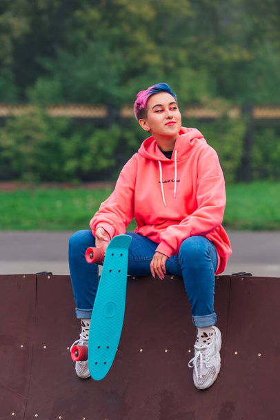 Portrait of a trendy pretty young girl with short colored hair and nose piercing dressed in pink hoodie and jeans sitting in skate park with her blue plastic skateboard. - Foto, Imagem