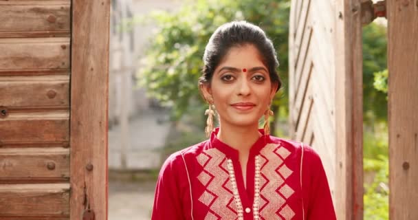Portrét hinduistické krásné mladé ženy s červenou tečkou na čele a v tradičním outfitu stojící venku v dřevěných dveřích a usmívající se. Okouzlující, radostná modelka v Indii. Hezká dívka v červených šatech. - Záběry, video