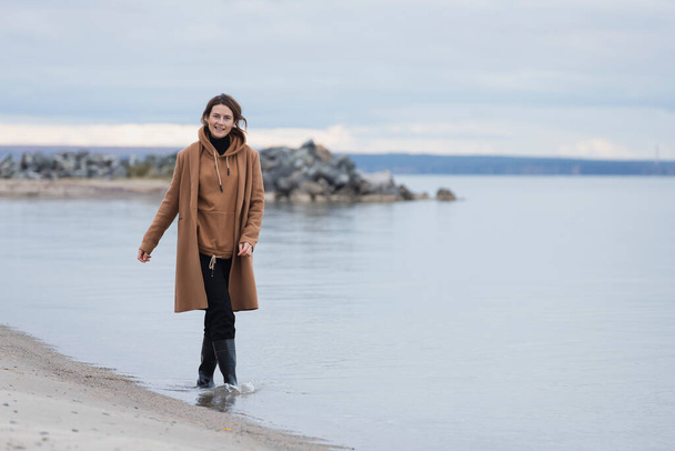 Portrait of a beautiful young model  in  a beige wool coat  enjoy day, on background  sea in  sunny autumn day. The concept of the unity of women and nature, peaceful mood - Photo, Image