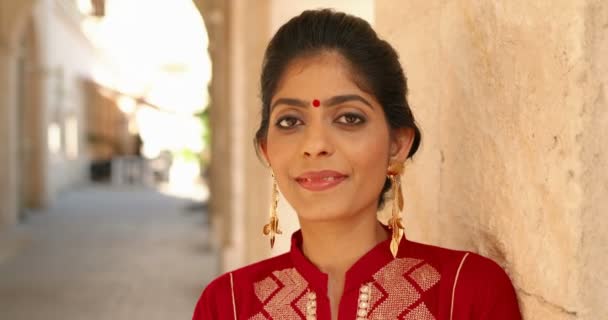 Primer plano de la joven hindú hermosa mujer con punto en la frente y en traje tradicional rojo, apoyado en la pared y sonriendo a la cámara. Retrato de chica alegre sonriente bastante encantadora de la India. - Metraje, vídeo