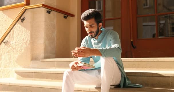 Schöner junger Hindu-Mann in traditioneller Kleidung sitzt auf Stufen im Freien und liest Notizen in Notizbüchern. Männchen aus Indien überprüfen Planer. Tagesplanung. Tagebuch. - Filmmaterial, Video