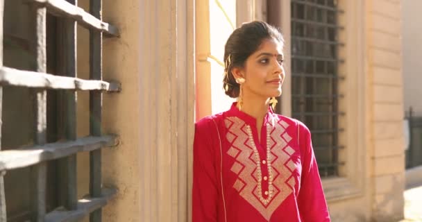 Portret van Hindoe jonge mooie vrouw met stip op het voorhoofd en in rode traditionele outfit draaien gezicht en glimlachen naar de camera. Buiten. Behoorlijk charmant gelukkig meisje uit India aan de muur op straat. - Video