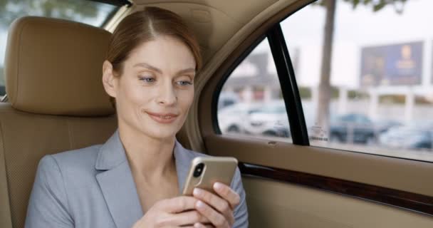 Close-up van jonge mooie blanke vrouw zittend op de achterbank van dure auto, glimlachend en sms 'end op smartphone. Vrij succesvolle zakenvrouw afluisteren en scrollen op mobiele telefoon. - Video