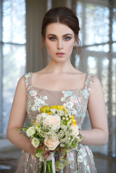 Portrait of a cute girl bride with a bouquet in her hands indoors on a window background. - Φωτογραφία, εικόνα