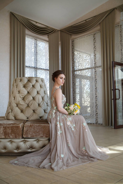 Lovely girl bride in a long flowing dress with a bouquet in her hands sitting in a chic interior. - Foto, Imagem