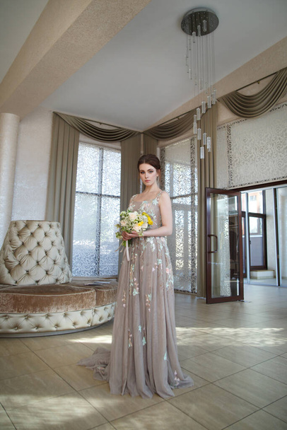 Lovely girl bride in a long flowing dress with a bouquet in her hands sitting in a chic interior. - Фото, зображення