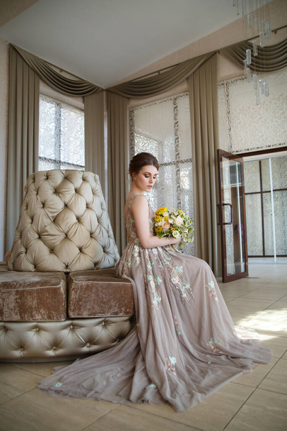 Lovely girl bride in a long flowing dress with a bouquet in her hands sitting in a chic interior. - Zdjęcie, obraz