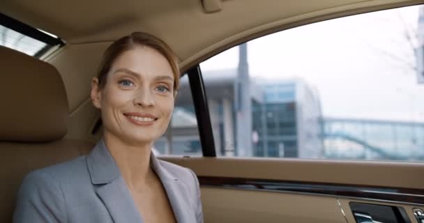 背部座席に高価な車に座っている間、若い白人の美しい実業家の肖像画は、カメラに顔を回し、誠実に笑みを浮かべている。笑顔でビジネススタイルの女性は午前中に仕事に行く - 映像、動画