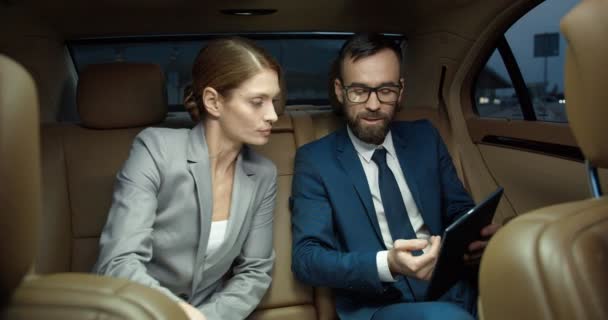 Καυκάσιος επιχειρηματίας που δείχνει κάτι στη γυναίκα επιχειρηματία στο tablet ενώ κάθονται μαζί στο πίσω κάθισμα του αυτοκινήτου και μιλάνε για δουλειές. Ζευγάρι εταίρων συζητούν εργασία και έργο. - Πλάνα, βίντεο
