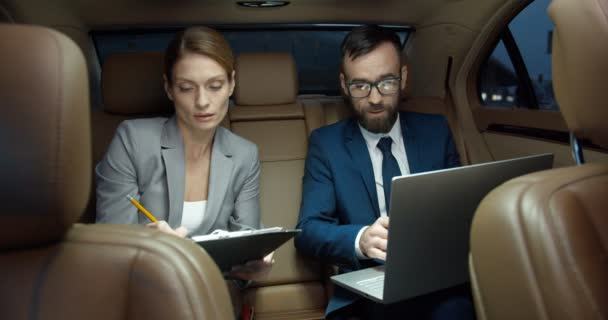 Άνδρες και γυναίκες Καυκάσιοι συνεργάτες κάθονται στο πίσω κάθισμα του αυτοκινήτου και εργάζονται μαζί με φορητό υπολογιστή και συμβόλαιο. Επιχειρηματίας με βοηθό σχεδιασμού εργασιών και ελέγχου εγγράφων. Κύριοι συνάδελφοι. - Πλάνα, βίντεο