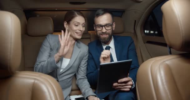白人のビジネスマンやビジネスマンが車の後部座席に座って、タブレットデバイスのウェブカメラを介してビデオチャットで話しています。ビジネスパートナーのカップルが笑顔とビデオ撮影。ビデオ通話。オンラインチャット - 映像、動画