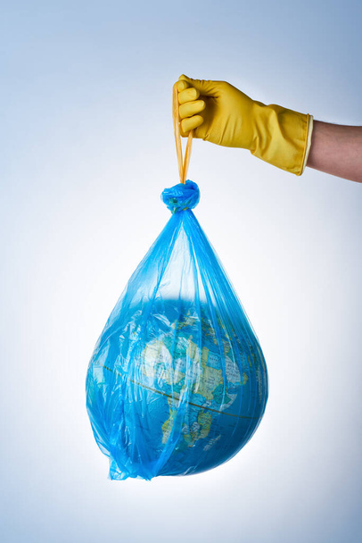 The concept of protecting the world from plastic waste, fighting environmental pollution, a hand in a yellow rubber glove holds a blue garbage bag with a globe on a light background. No plastic bags - Zdjęcie, obraz