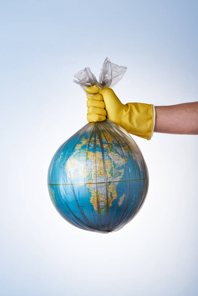 The concept of protecting the world from plastic waste, fighting environmental pollution, a hand in a yellow rubber glove holds a black garbage bag with a globe on a light background. No plastic bags - Fotó, kép