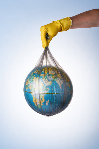 The concept of protecting the world from plastic waste, fighting environmental pollution, a hand in a yellow rubber glove holds a black garbage bag with a globe on a light background. No plastic bags - Фото, изображение