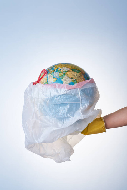 Ajatus maailman suojelemisesta muovijätteeltä, ympäristön pilaantumisen torjunta, käsi keltaisessa kumihanskassa pitää valkoista roskapussia, jossa on maapallo kevyellä taustalla. Ei muovipusseja - Valokuva, kuva