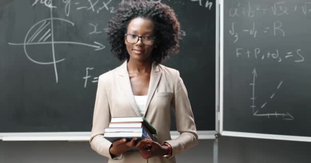 Gözlüklü genç Afrikalı Amerikalı kadın öğretmenin portresi. Sınıfta kameraya bakıyor ve ders kitaplarını tutuyor. Arkaplanda formüller olan karatahta. Okul konsepti. Kadınların elindeki kitaplar.. - Video, Çekim