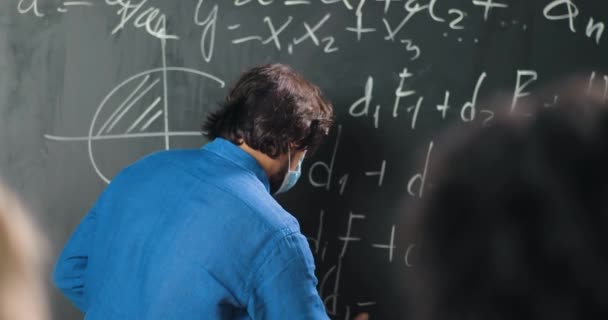 Kaukázusi férfi tanár orvosi maszkban az iskolában képleteket és matematikai törvényeket ír a táblán. Iskolai koncepció. Férfi előadó szemüvegben, aki a fizika törvényeit magyarázza. A koronavírus fogalma. - Felvétel, videó