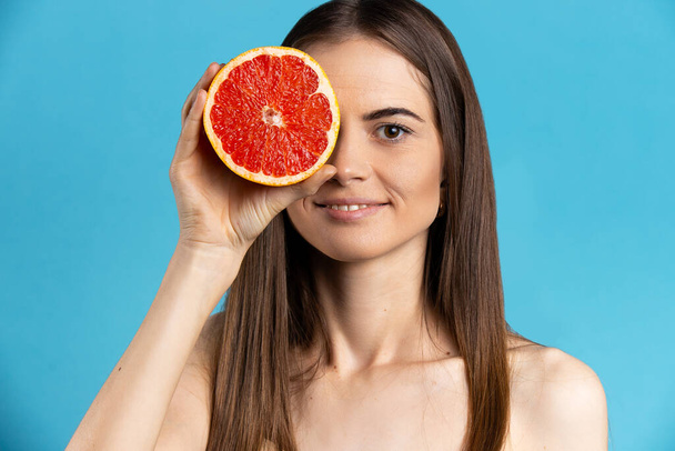Natural organic raw fresh food concept. Smiling, attractive girl holding half of grapefruit on blue isolated background - Zdjęcie, obraz