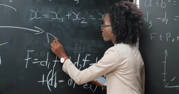 Terug blik op Afro-Amerikaanse jonge vrouw leraar op school schrijven formules en wiskunde wetten op schoolbord. Schoolconcept. Vrouwelijke docent in glazen die natuurkunde wetten uitlegt. Achterzijde. - Video