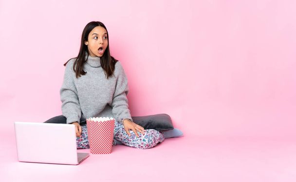Jovem mulher de raça mista comendo pipocas enquanto assiste a um filme no laptop fazendo gesto surpresa enquanto olha para o lado - Foto, Imagem