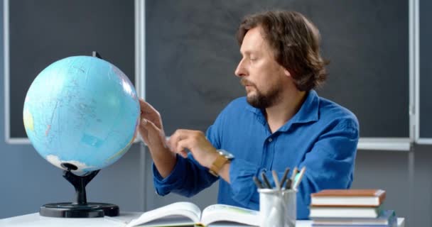 Egy fehér férfi tanár portréja ül az asztalnál, és a földgömböt vizsgálja az osztályban. Földrajzot tanít az osztályteremben. Online lecke. A világról szóló témát magyarázó előadó. - Felvétel, videó