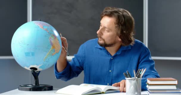 Retrato de un maestro caucásico sentado a la mesa y examinando el globo en clase, anotando en un libro de texto con lápiz. Hombre leyendo libro de geografía en el aula. Lección en línea. Profesor preparándose para la lección - Imágenes, Vídeo