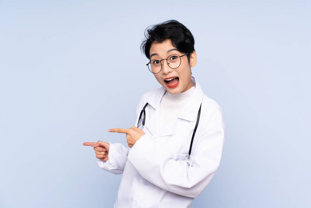 Doctor Asian woman over isolated blue background surprised and pointing side - Photo, image