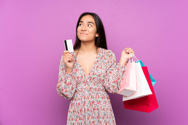 Young Indian woman isolated on purple background holding shopping bags and a credit card and thinking - Photo, Image