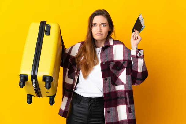 Νεαρή Ιρλανδή γυναίκα απομονωμένη σε κίτρινο φόντο δυστυχισμένη σε διακοπές με βαλίτσα και διαβατήριο - Φωτογραφία, εικόνα