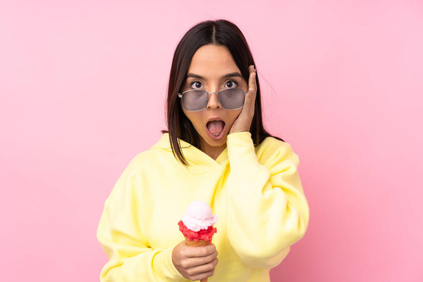 Giovane ragazza bruna che tiene un gelato alla cornetta su sfondo rosa isolato con sorpresa e espressione facciale scioccata - Foto, immagini