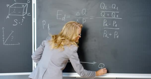 Visszapillantás a kaukázusi női előadóról, aki matematikai vagy fizika képleteket ír és grafikát rajzol krétákkal a táblán. Női tanár dolgozik az iskolában. Matematikai lecke koncepció. Hátsó rész. - Felvétel, videó