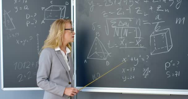 Iloinen valkoihoinen kaunis naisopettaja laseissa seisoo liitutaulussa kaavoilla, grafiikalla ja laeilla ja selittää osoittimella. Matematiikan opettaja koulussa tai korkeakoulussa. - Materiaali, video
