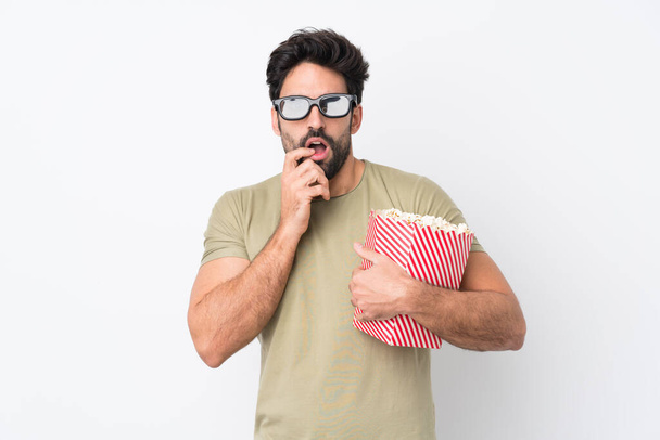 Young handsome man with beard over isolated white background with 3d glasses and holding a big bucket of popcorns - Foto, imagen