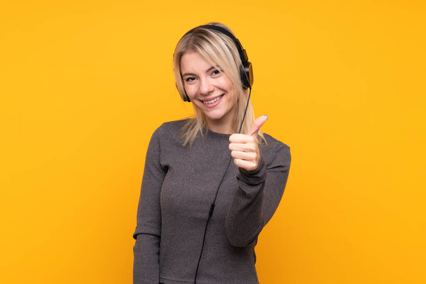 Young blonde woman over isolated yellow background listening music and with thumb up - Foto, afbeelding