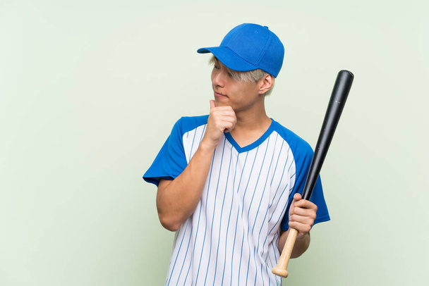 Молодой азиат, играющий в бейсбол на изолированном зеленом фоне, думает об идее и смотрит в сторону - Фото, изображение