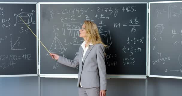 Blanke mooie vrouwelijke leraar in glazen staande op schoolbord met formules, afbeeldingen en wetten en uit te leggen met pointer. Docent wiskunde praat met camera 's op school. Wiskundeles. - Video