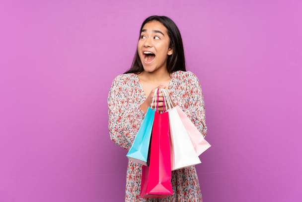 Young Indian woman isolated on purple background holding shopping bags and surprised - Photo, Image