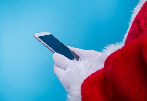 Santa Claus holding mobile phone ready for Christmas time on blue  background. The season, winter, holiday, celebration, gift concept - Photo, Image