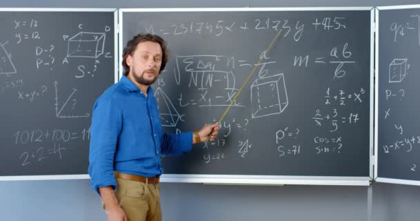 Valkoihoinen miespuolinen matematiikan opettaja seisoo taululla kaavojen, grafiikan ja lakien kanssa ja selittää fysiikkaa osoittimella. Matematiikan opettaja koulussa tai korkeakoulussa. Opetussuunnitelma. - Materiaali, video
