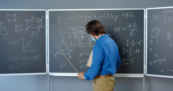 Белый красавец-учитель в медицинской маске, стоящий у доски с формулами, графикой и законами и объясняющий указателем. Преподаватель математики в школе или колледже. Коронавирусная концепция. - Кадры, видео