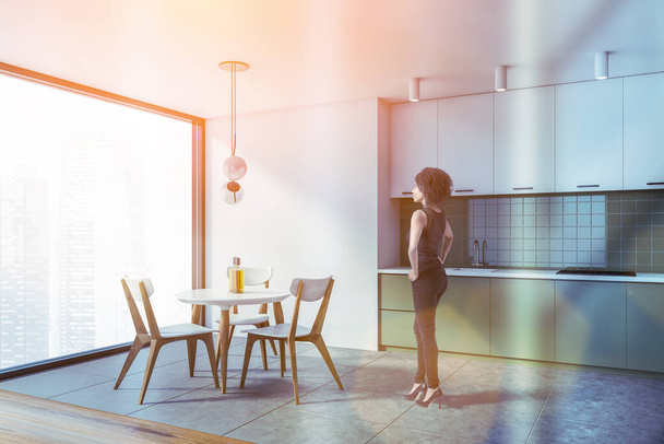 Beautiful young African American woman standing in modern kitchen with white walls, concrete floor and green cabinets. Round dining table. Toned image - Foto, imagen
