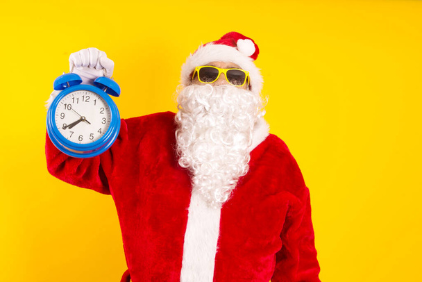 Santa Claus holding alarm clock in yellow background - Фото, изображение