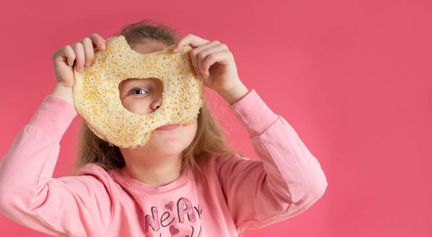 Μικρό χαριτωμένο κορίτσι κρατά μια τηγανίτα και κοιτάζει μέσα από μια τρύπα σε αυτό, σε ένα ροζ φόντο. Ωραίο πρωινό. καρναβάλι ανοιξιάτικο - Φωτογραφία, εικόνα